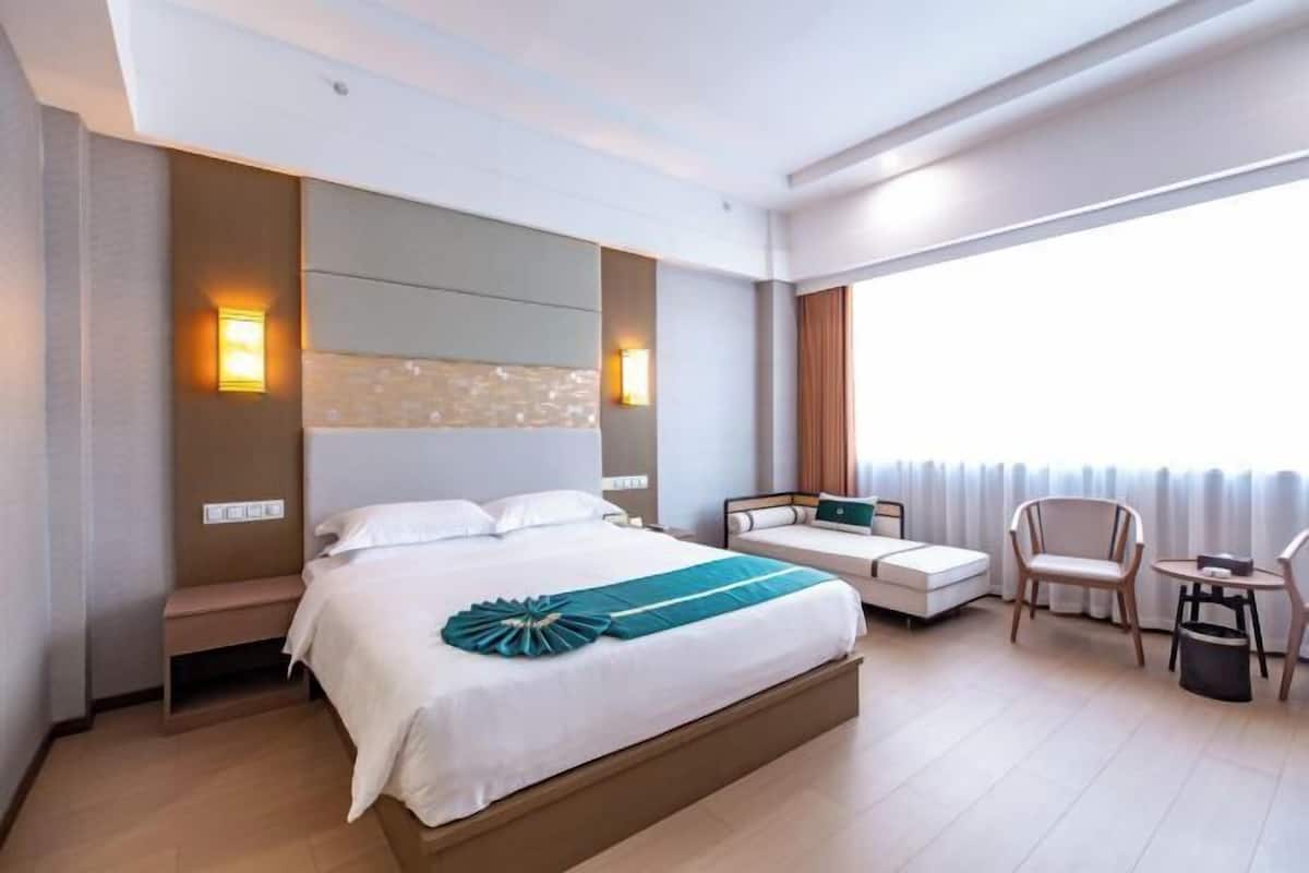 Una habitación de hotel moderna con una cama grande, mesitas de noche, una zona de estar con una silla y una pequeña mesa, y vistas al exterior.