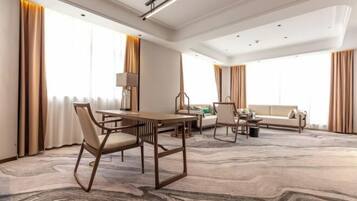 A spacious hotel room with a large dining table, a sofa, and a coffee table. There are large windows with curtains, a ceiling light, and a carpeted floor.