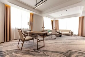 Une chambre d’hôtel spacieuse, dotée d’une grande table à manger, d’un canapé et d’une table basse. De grandes fenêtres sont agrémentées de rideaux, et la pièce est éclairée par un lustre au plafond. Le sol est recouvert d’une moquette.