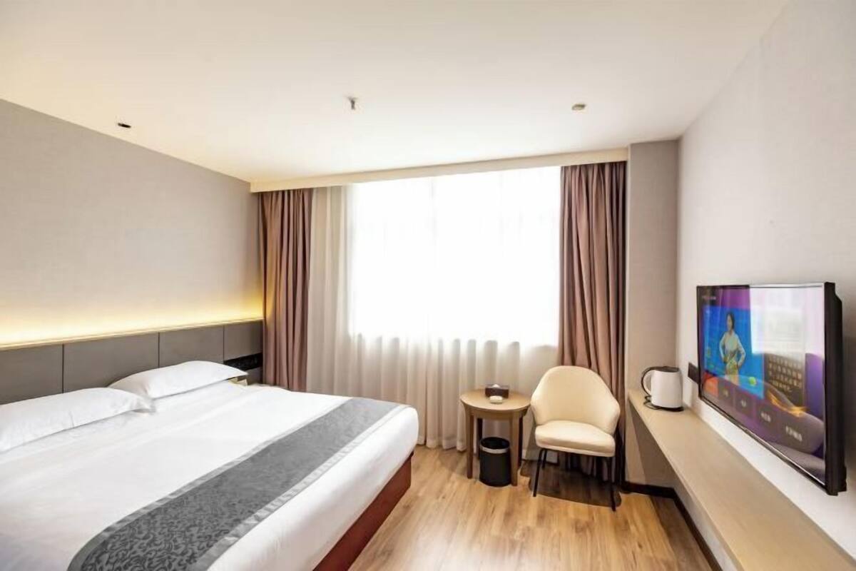 Una habitación de hotel con cama, televisión, sillón, mesita y ventana con cortinas.