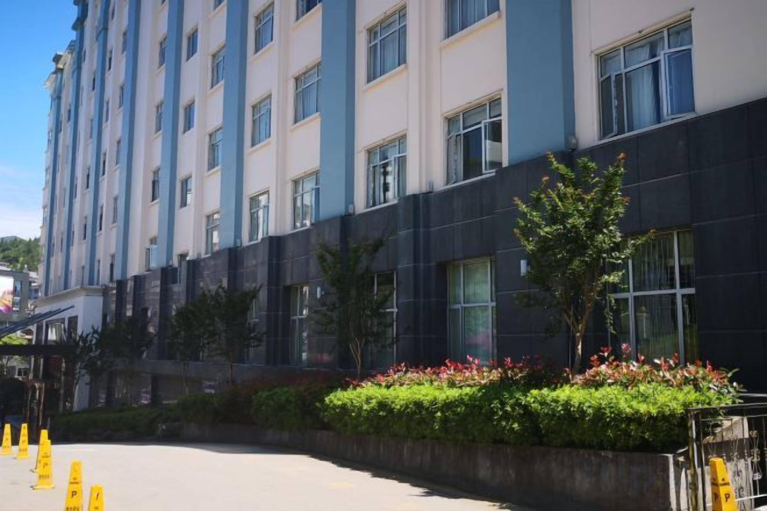 A multi-story hotel with a modern facade, a landscaped garden area with a small tree, and a clear sky.