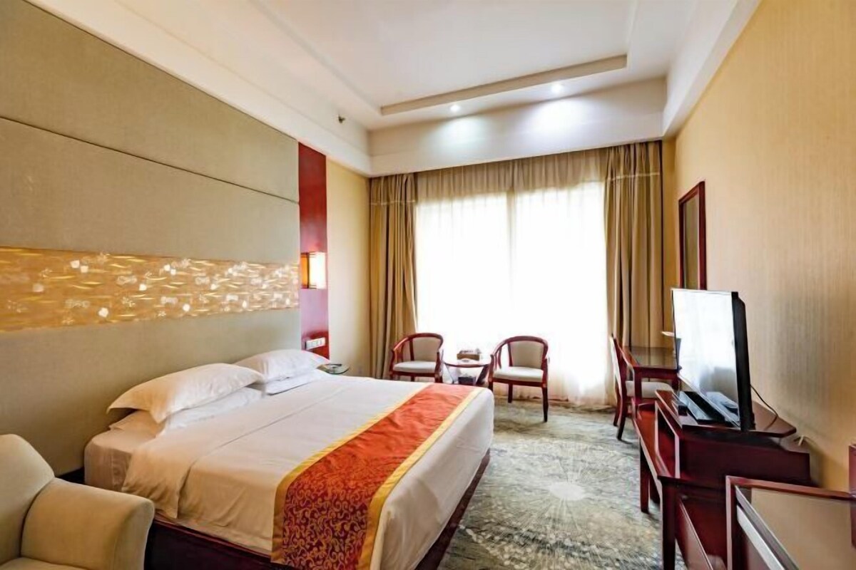 Une chambre d’hôtel avec un grand lit, un bureau avec une télévision, deux chaises et une fenêtre avec des rideaux.