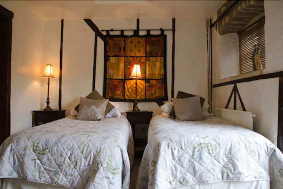 A room with two beds, each with a quilted bedspread, two bedside lamps, and a decorative stained glass window above the beds.