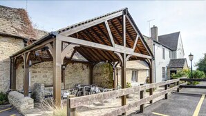 A covered outdoor seating area with tables and chairs, adjacent to a stone building with a pitched roof.