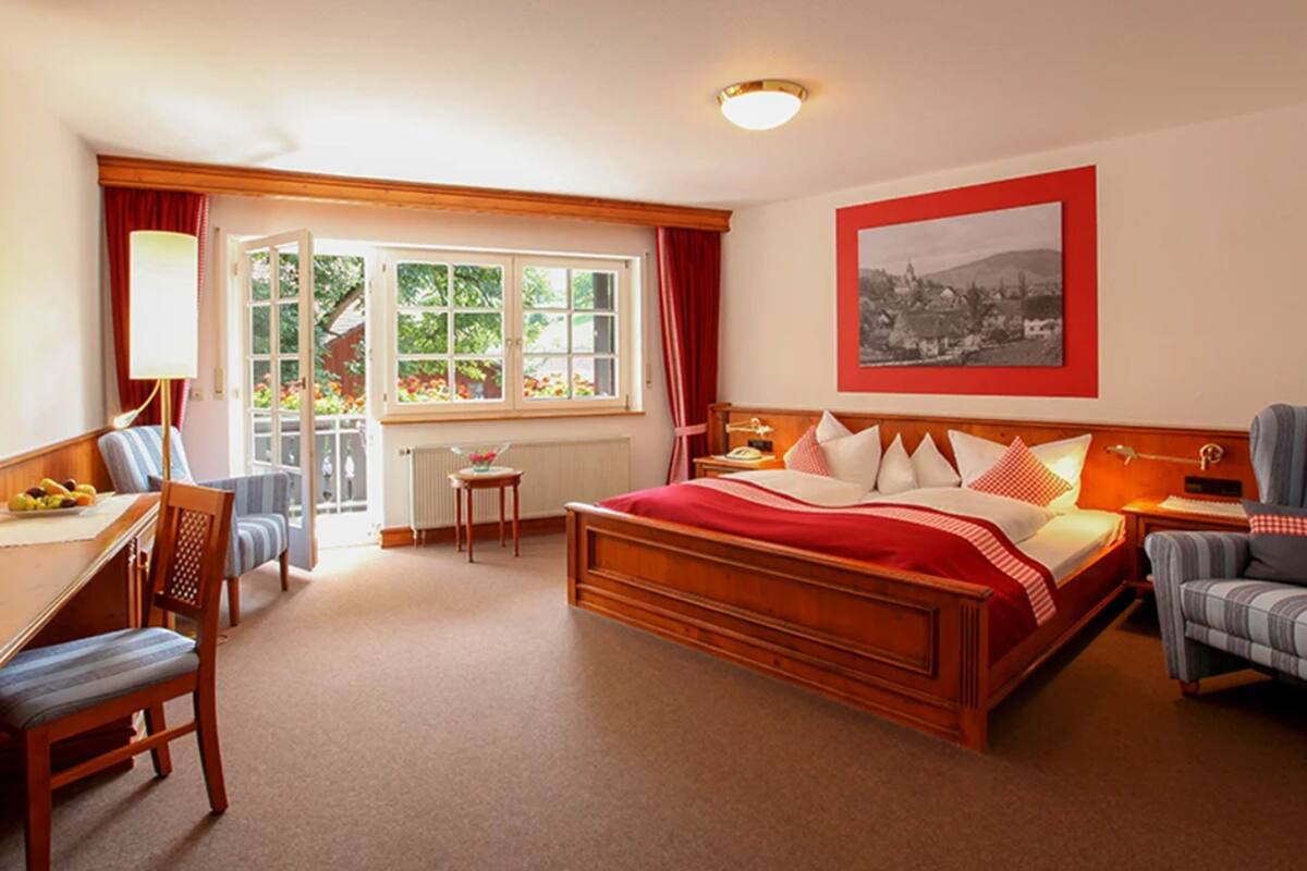 Ein Hotelzimmer mit einem hölzernen Bett, einem Schreibtisch mit Stuhl, einem kleinen Tisch und einem Fenster mit Vorhängen.