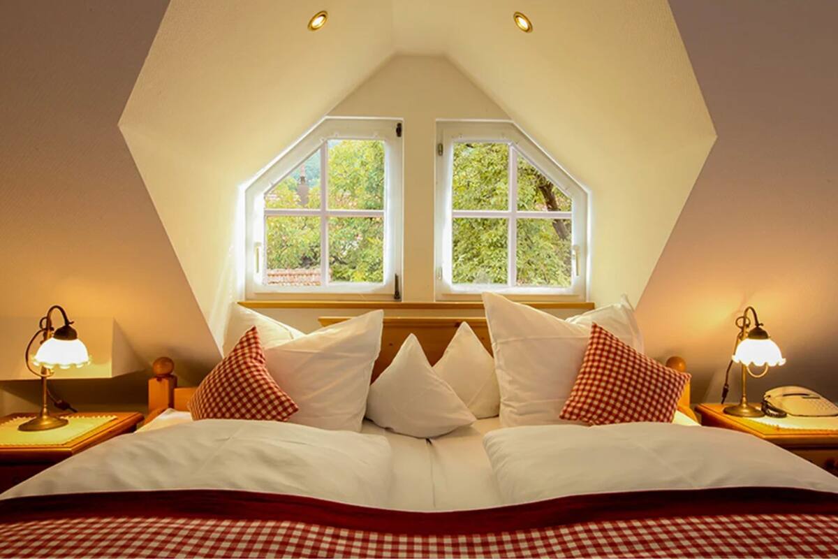 Ein Schlafzimmer mit geneigter Decke, zwei Fenstern mit weißen Rahmen, einem Bett mit weißer Bettwäsche und roten Karokissen sowie Nachttischlampen.