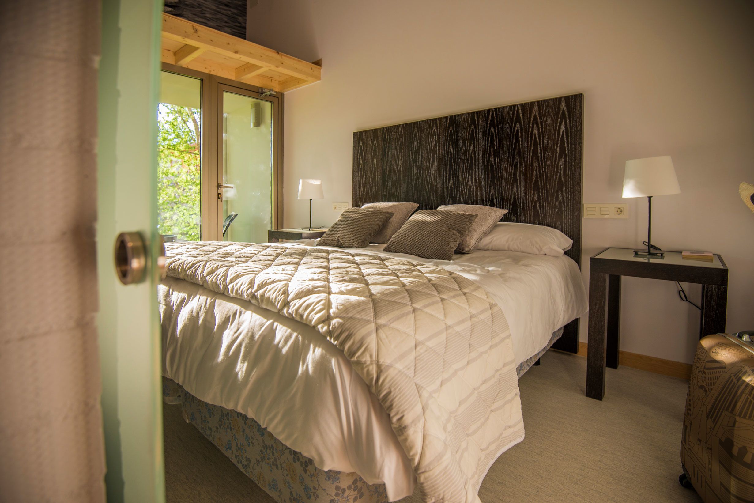 A bedroom with a large bed, two bedside tables with lamps, a wooden headboard, and a view of greenery outside.