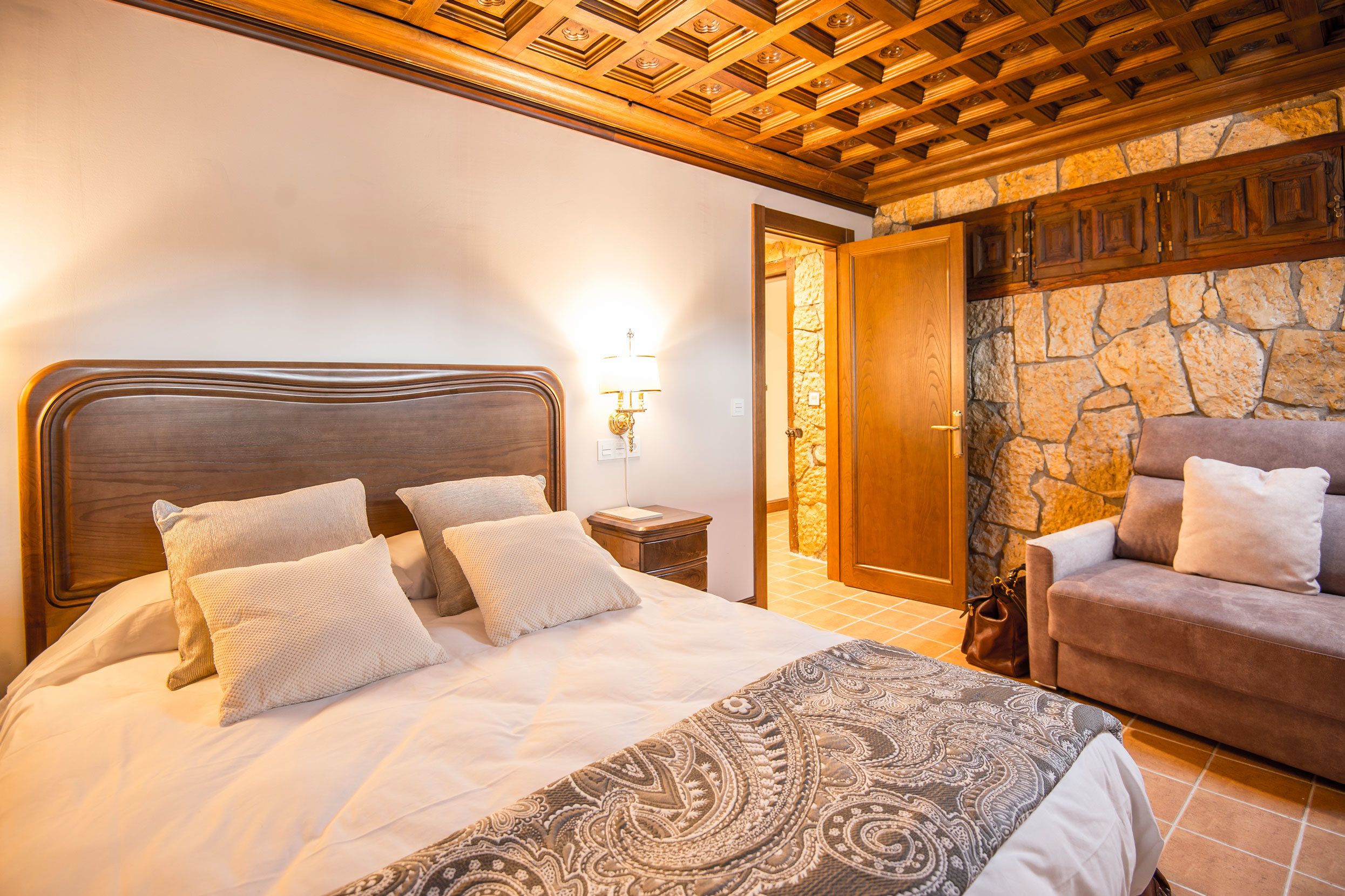 Ein helles Schlafzimmer mit einem großen Bett, einem Nachttisch, einer Lampe, einer Steinwand und einer Holztür.
