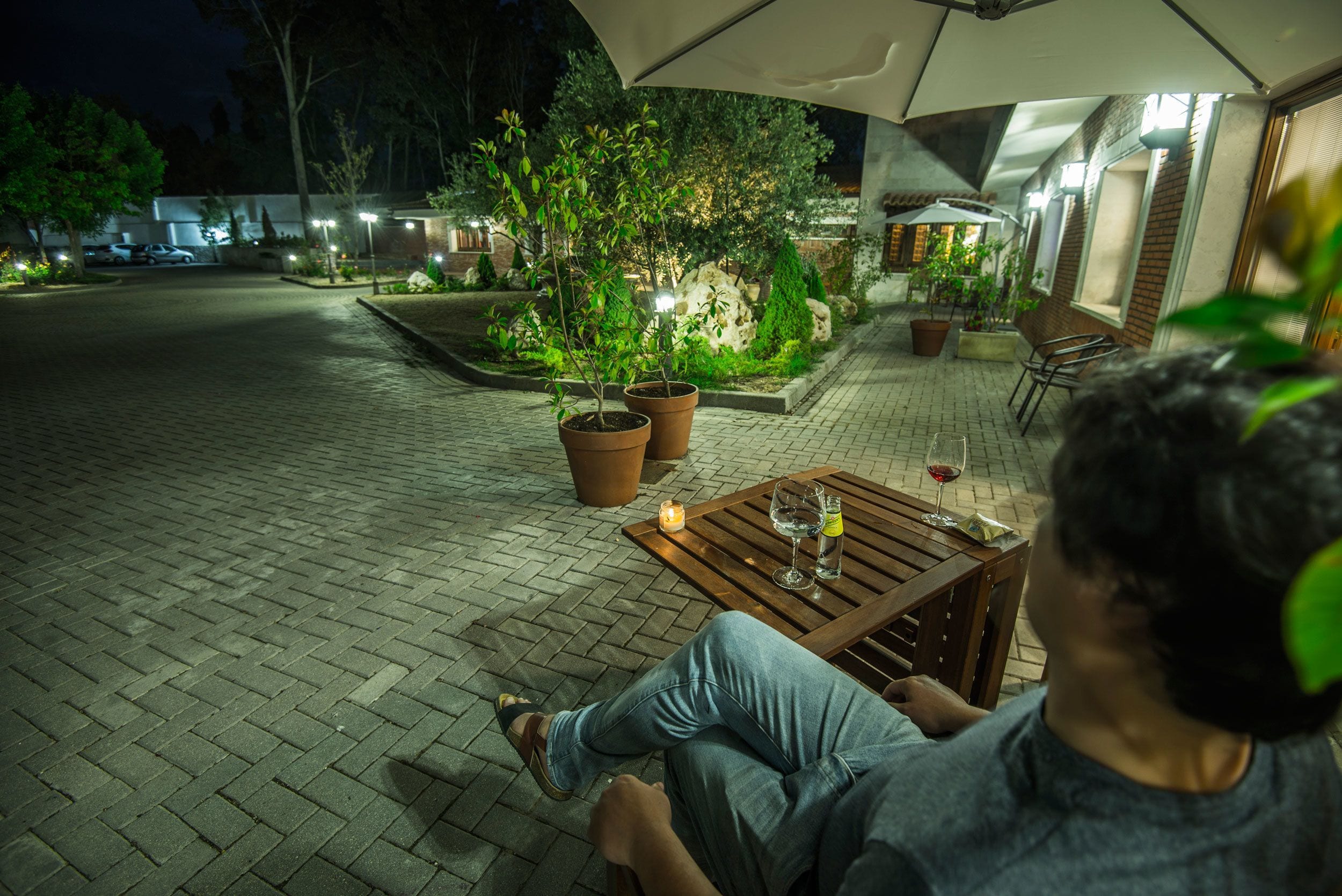 Une personne est assise à une table à l’extérieur, avec une bougie, sous un grand parasol, dans un espace patio bien éclairé, agrémenté de plantes en pot et doté d’un sol en briques.