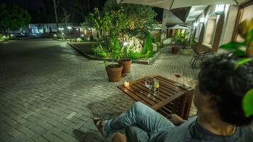 Une personne est assise à une table à l’extérieur, avec une bougie, sous un grand parasol, dans un espace patio bien éclairé, agrémenté de plantes en pot et doté d’un sol en briques.