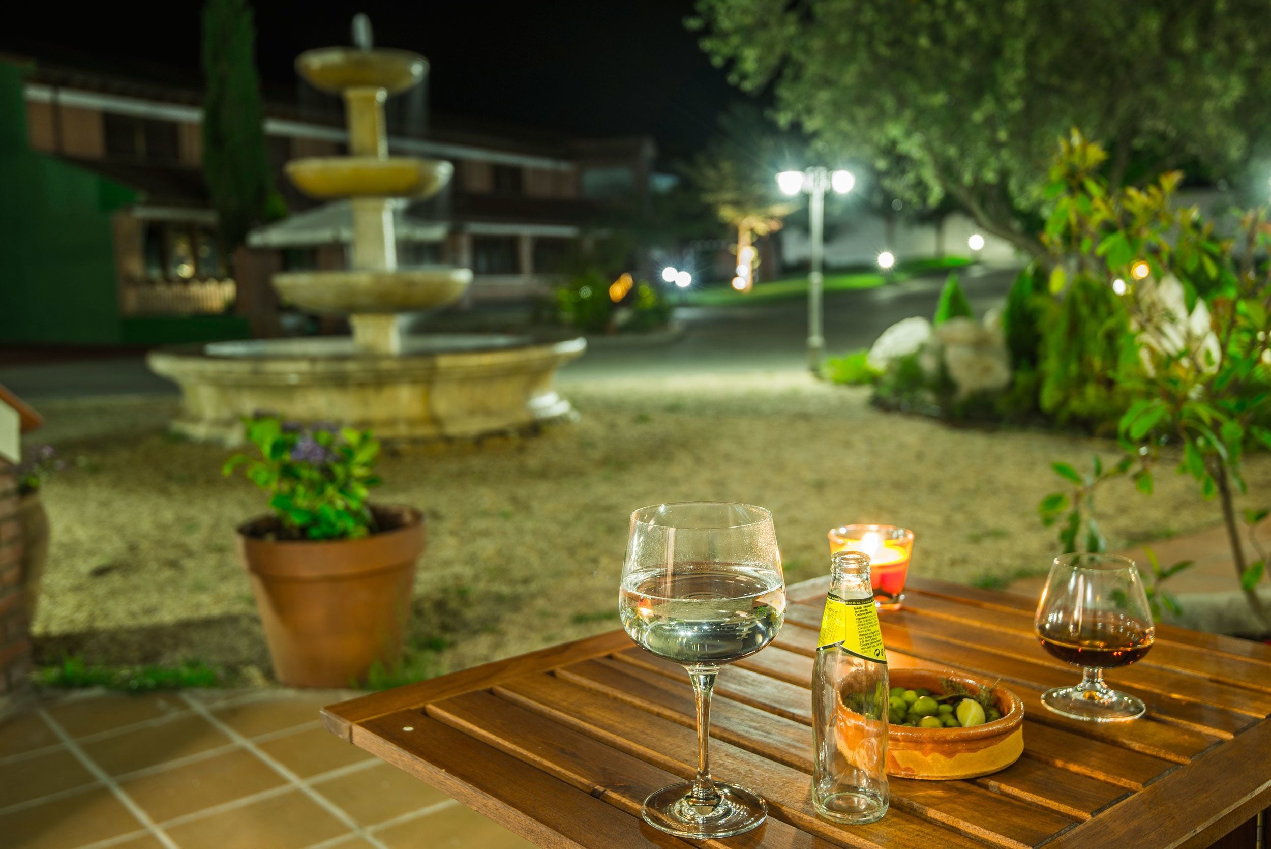 A wooden table with two wine glasses, a bottle, and a candle in an outdoor setting at night.