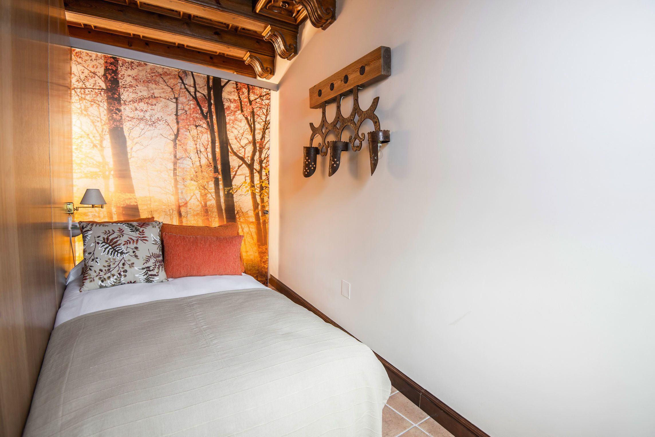 A bedroom with a bed, a nightstand, a wall-mounted light fixture, and a window with a view of trees.