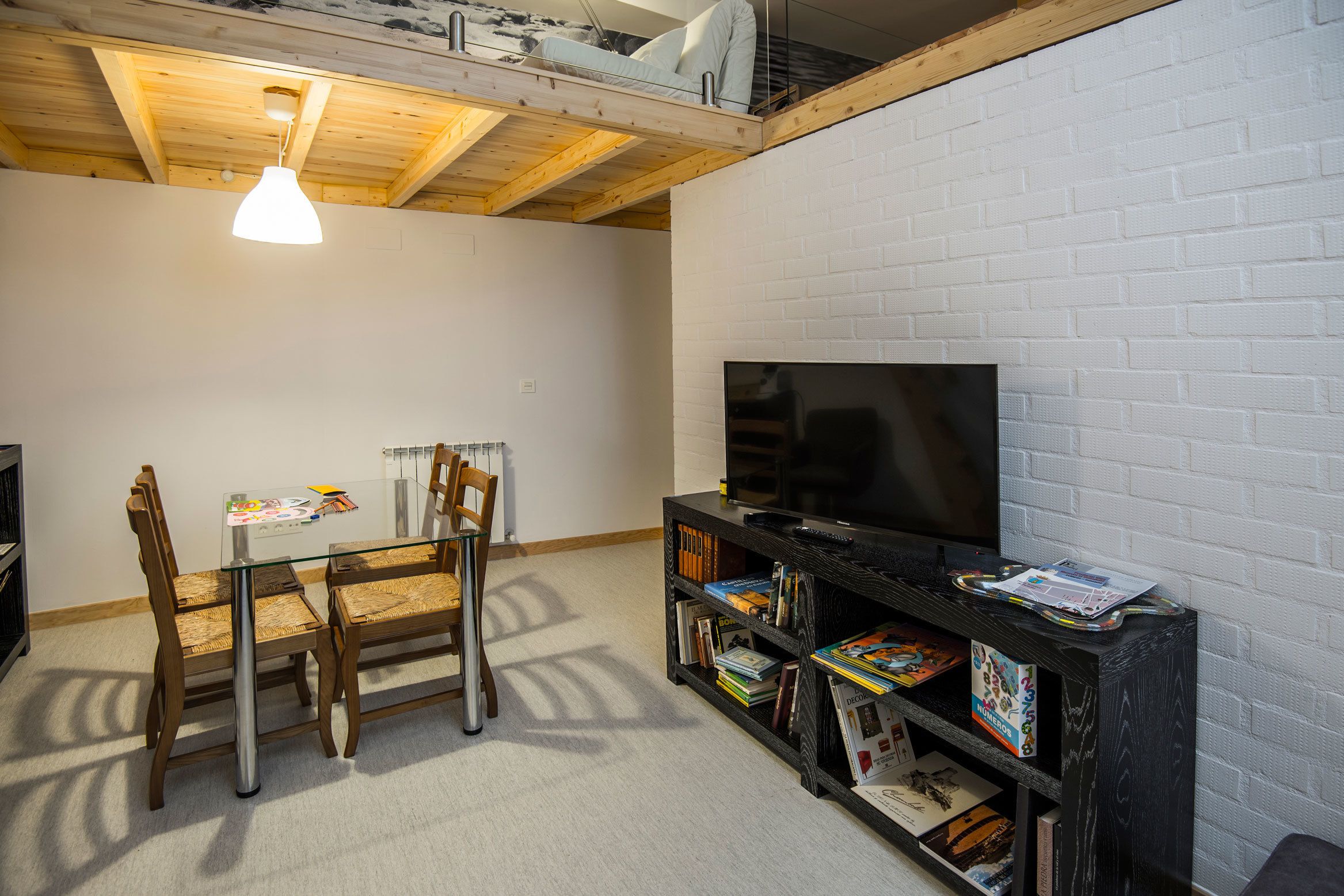 A modern living room with a glass table, wooden chairs, a black entertainment unit with books, and a TV.