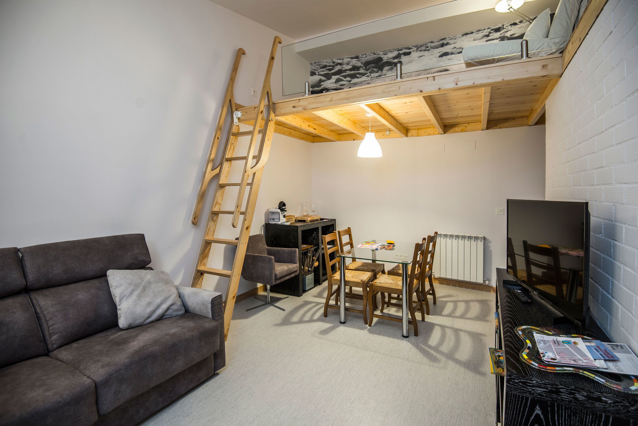A loft-style room with a bunk bed, a dining table with chairs, a sofa, a ladder, and a TV.