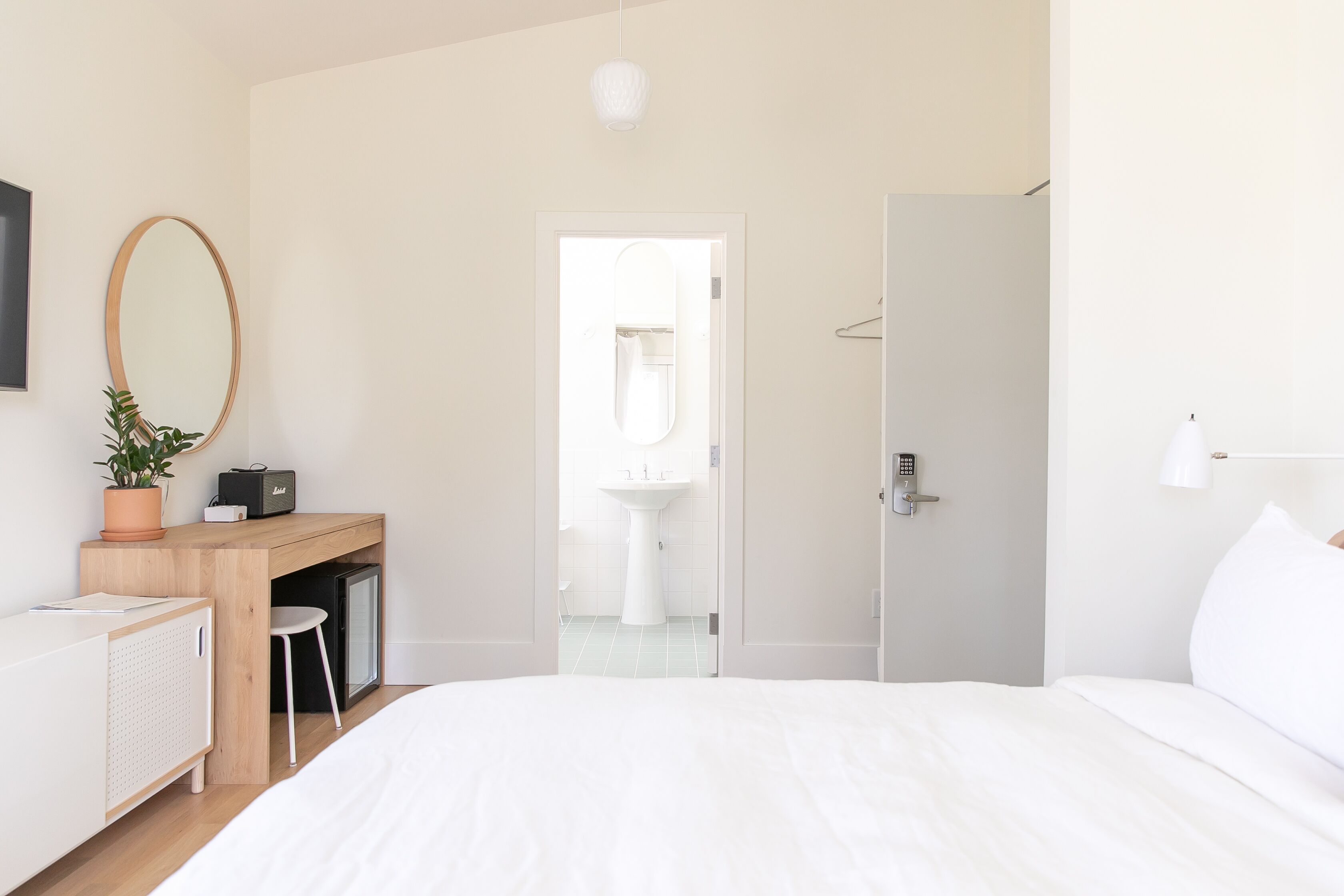 A modern hotel room with a bed, a desk with a computer, a potted plant, and a bathroom visible through an open door.