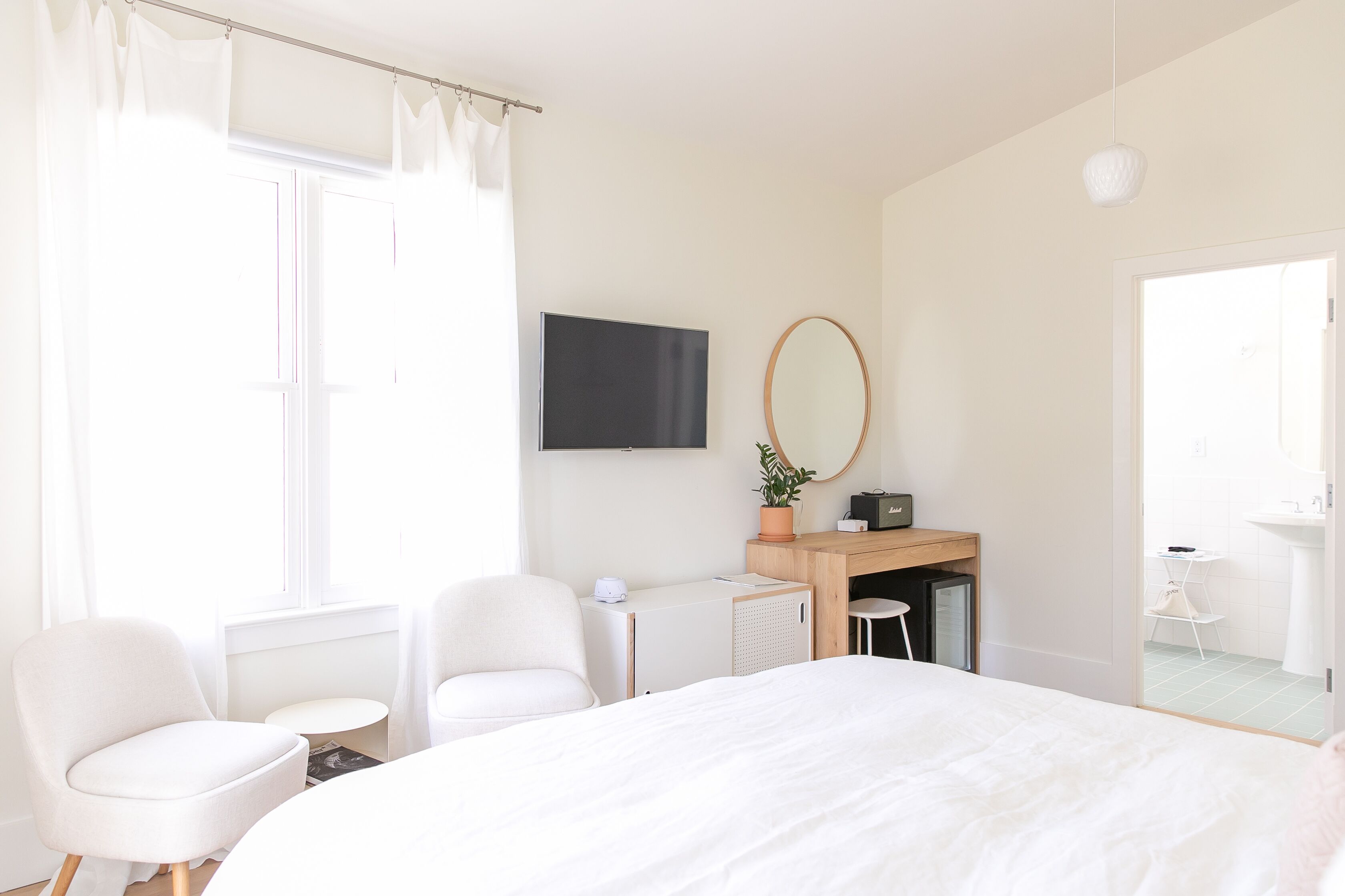 Habitación de hotel moderna con una cama grande, dos sillas blancas, un escritorio con una computadora, un televisor y un baño visible a través de una puerta abierta.