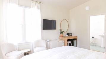 Habitación de hotel moderna con una cama grande, dos sillas blancas, un escritorio con una computadora, un televisor y un baño visible a través de una puerta abierta.