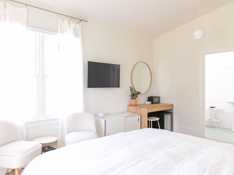 Habitación de hotel moderna con una cama grande, dos sillas blancas, un escritorio con una computadora, un televisor y un baño visible a través de una puerta abierta.