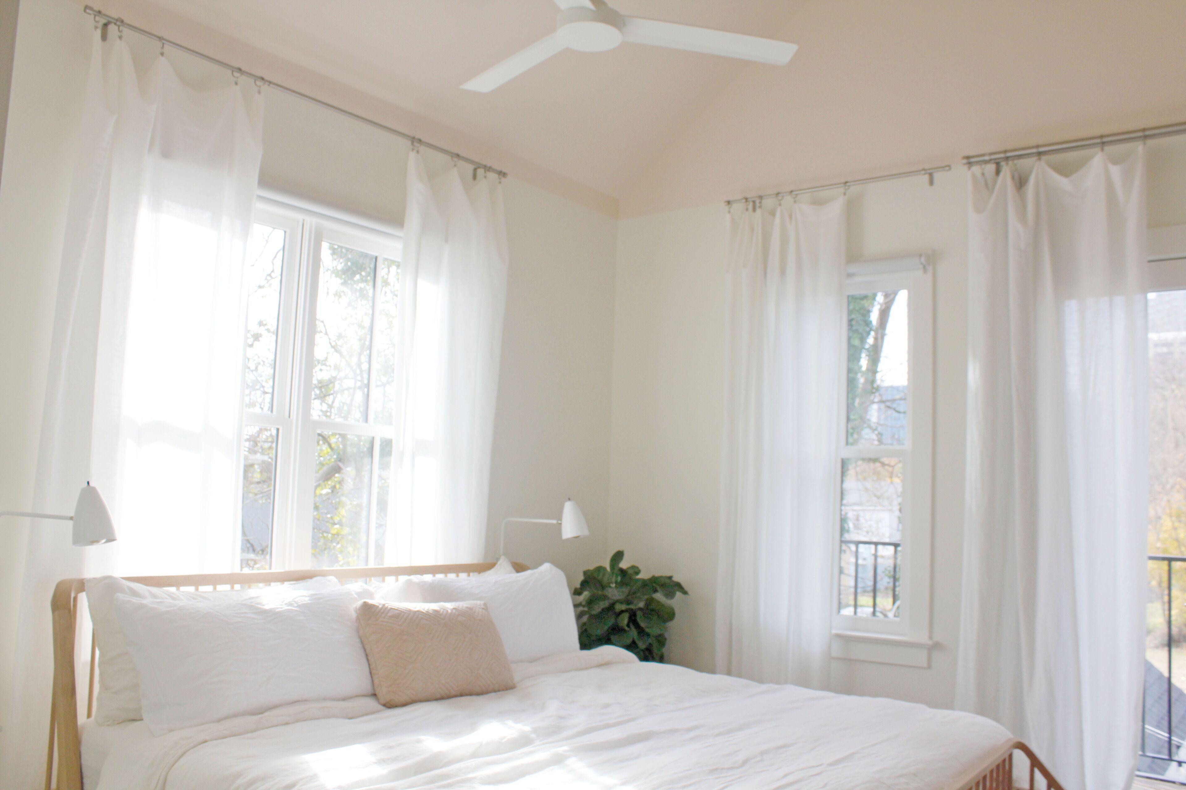 A bedroom with a bed, white curtains, a ceiling fan, and a window.