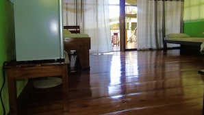 Una habitación con piso de madera, una cama, una silla y una mesa con refrigerador.