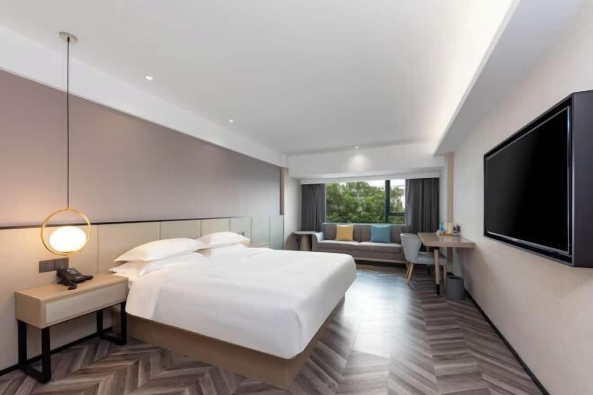 A hotel room with a bed, bedside table, lamp, TV, seating area, and a view of greenery outside.