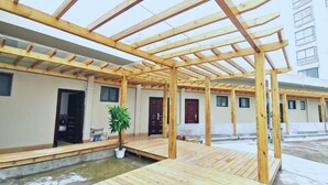 Ein überdachter Außenbereich mit einer Holz-Pergola, einem kleinen Wasserspiel und einem Topfpflanzen.