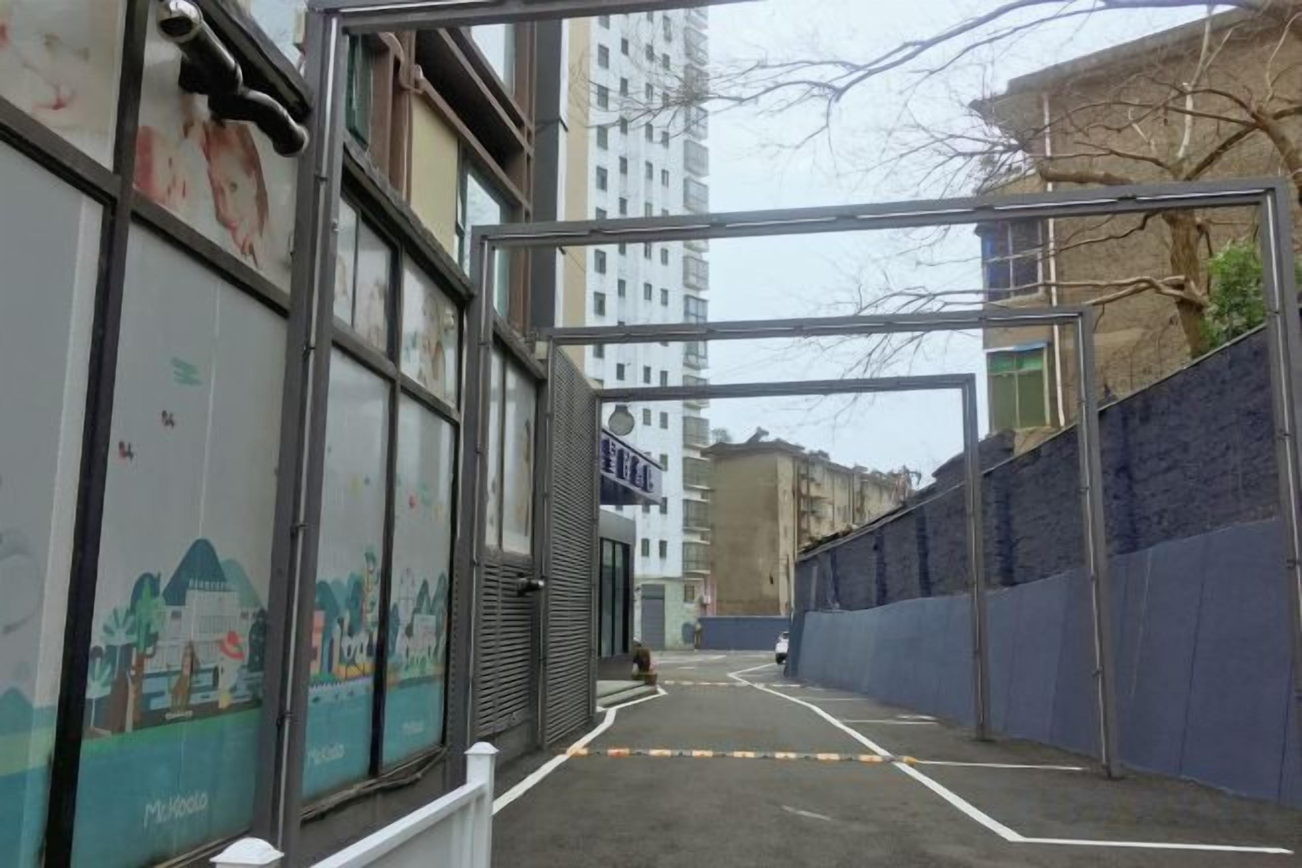 A narrow alleyway with a metal structure overhead, flanked by buildings with windows and a mural on one wall.