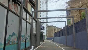 A narrow alleyway with a metal structure overhead, flanked by buildings with windows and a mural on one wall.