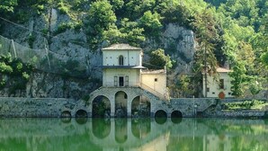 A stone bridge with arches leading to a building with a tiled roof, situated by a calm body of water with reflections. - Albergo Miramonti (Scanno)