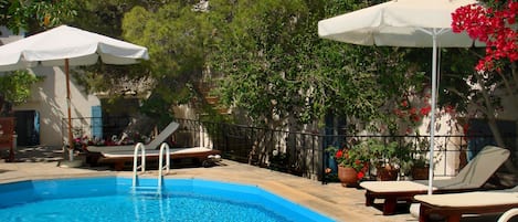 Una piscina con sillones de descanso y sombrillas, rodeada de plantas en macetas y árboles.