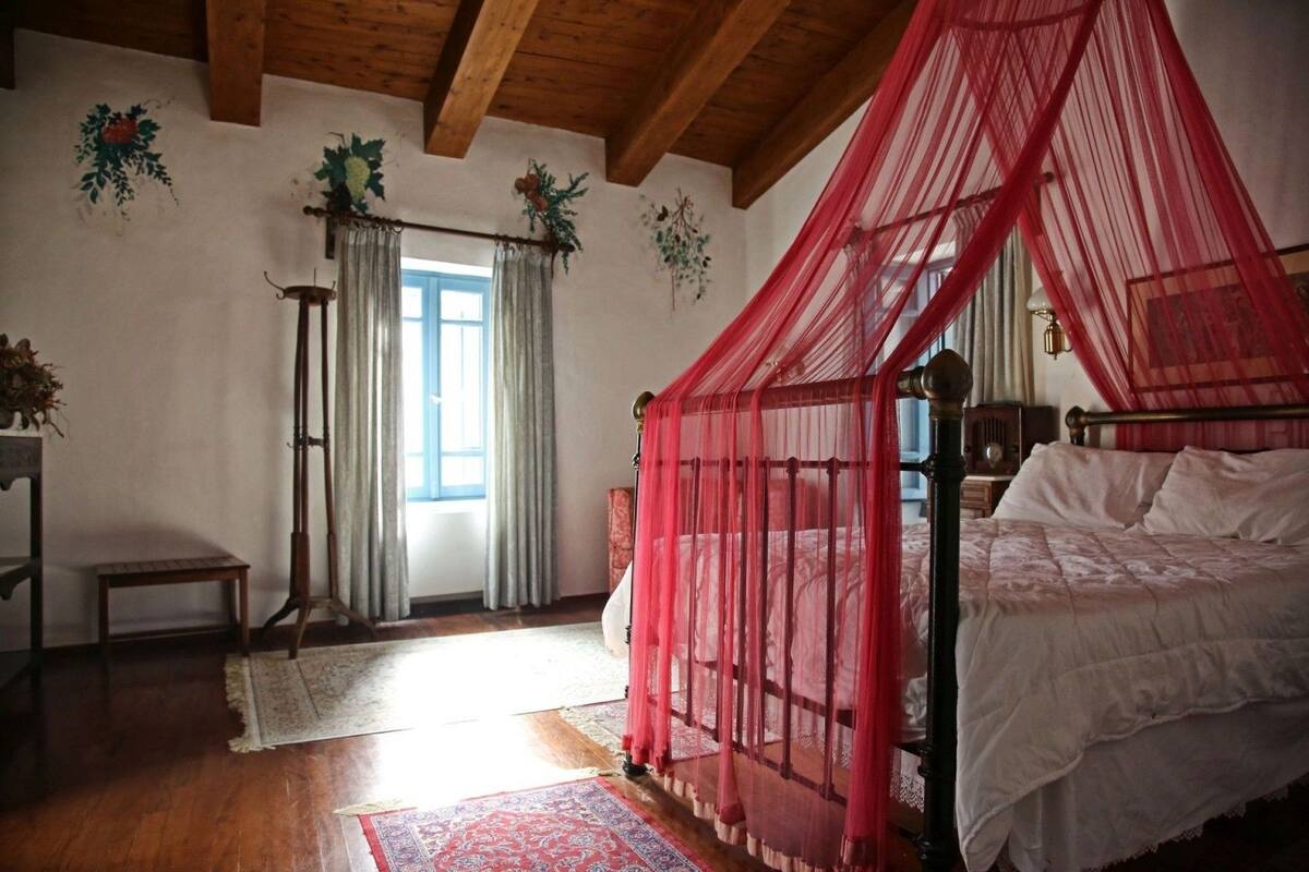 A bedroom with a four-poster bed, a wooden floor, a red canopy, and a window with curtains.