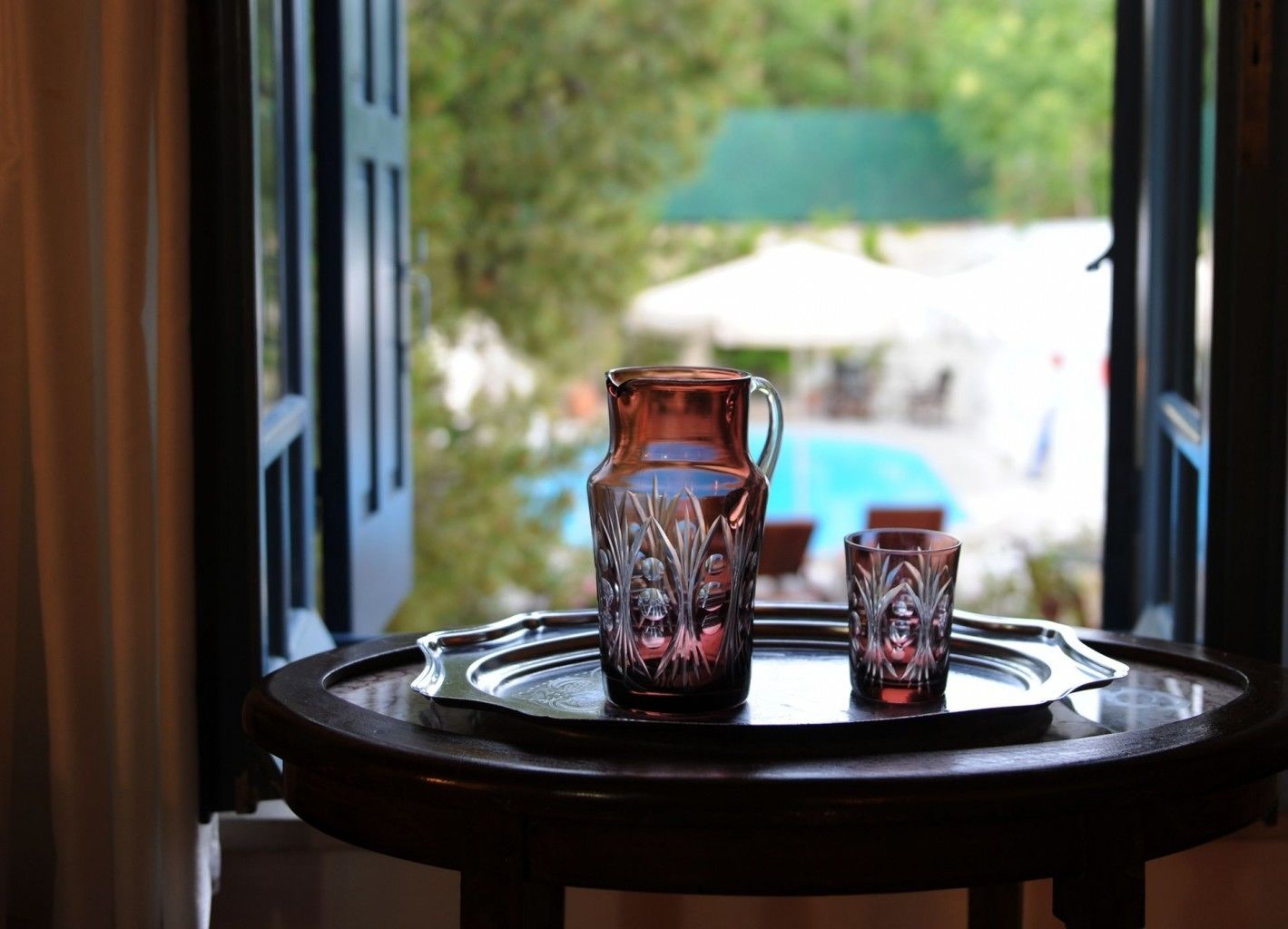 Una mesa con una jarrón y un vaso, frente a una ventana con vista a una piscina y follaje.