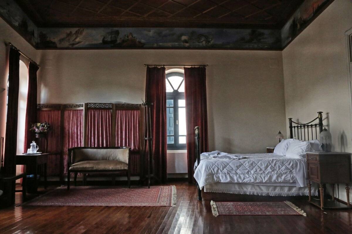 A traditional room with a bed, a chair, a table with a vase, and a window with curtains.