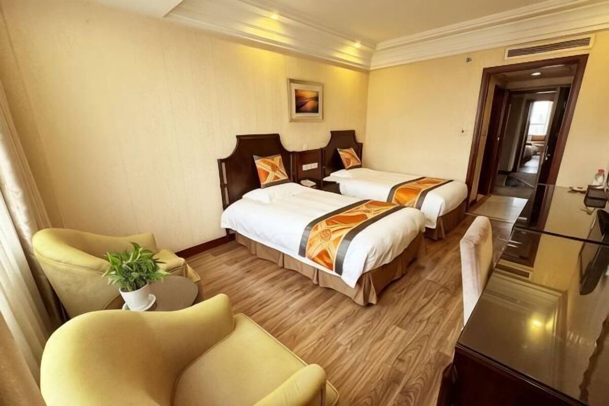 A hotel room with two beds, a desk, a chair, a small table with a plant, and a mirror.