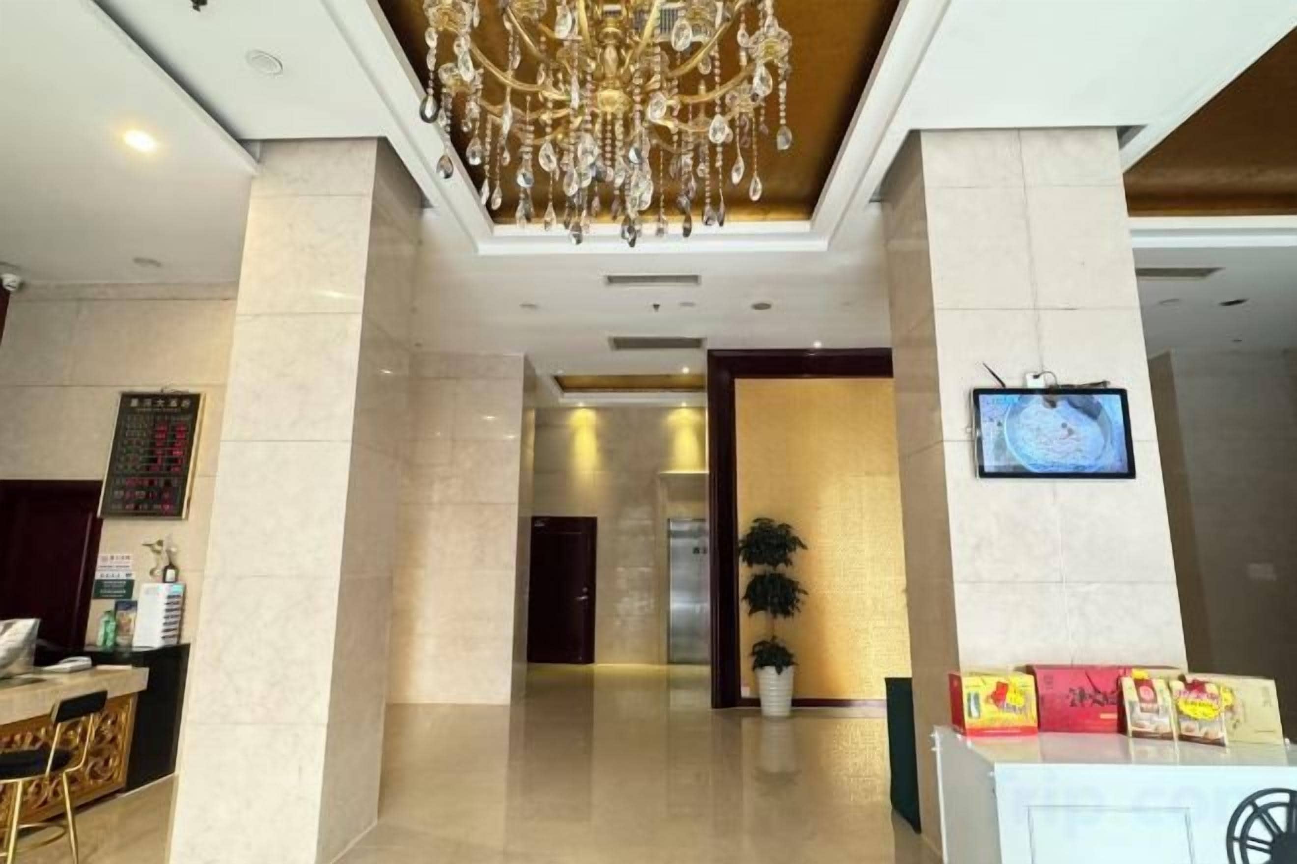 A hotel lobby with a chandelier, a reception desk, a television screen, and a potted plant.