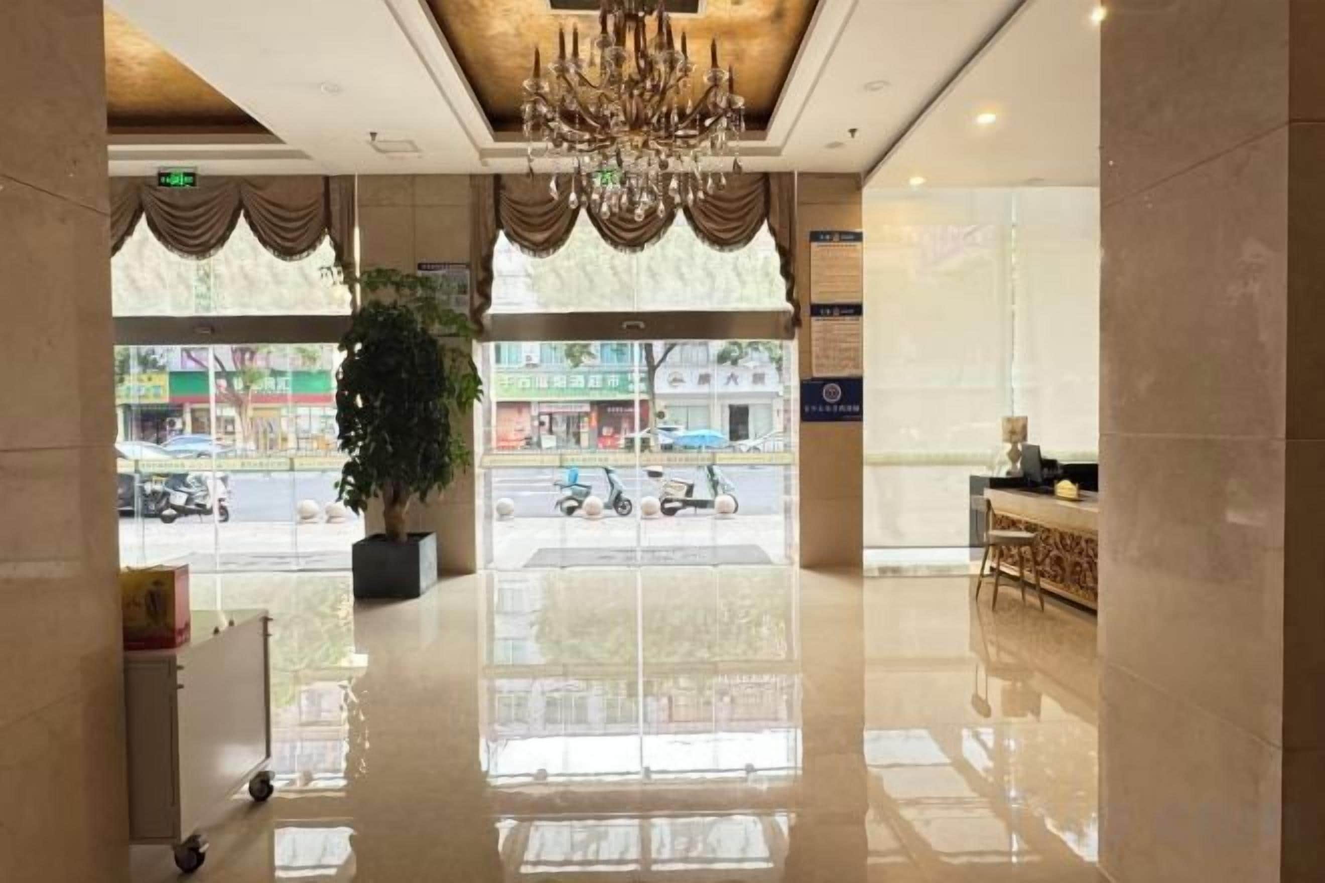 A hotel lobby with a chandelier, a reception desk, and a large window offering a view of the street.