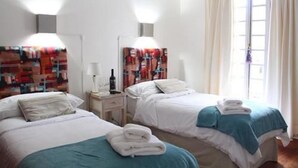 A hotel room with two beds, a nightstand with a lamp, a window with curtains, and a wall with a colorful abstract painting. - Chiara Lodge (Chacras de Coria)