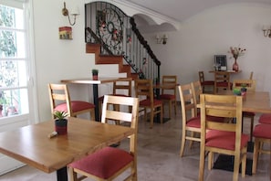A dining area with wooden tables and chairs, a staircase with decorative railings, and a view of the outside through a window.