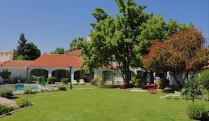 Una villa con techo de tejas rojas, piscina y un jardín bien cuidado con diversas plantas y un gran árbol con flores rojas.