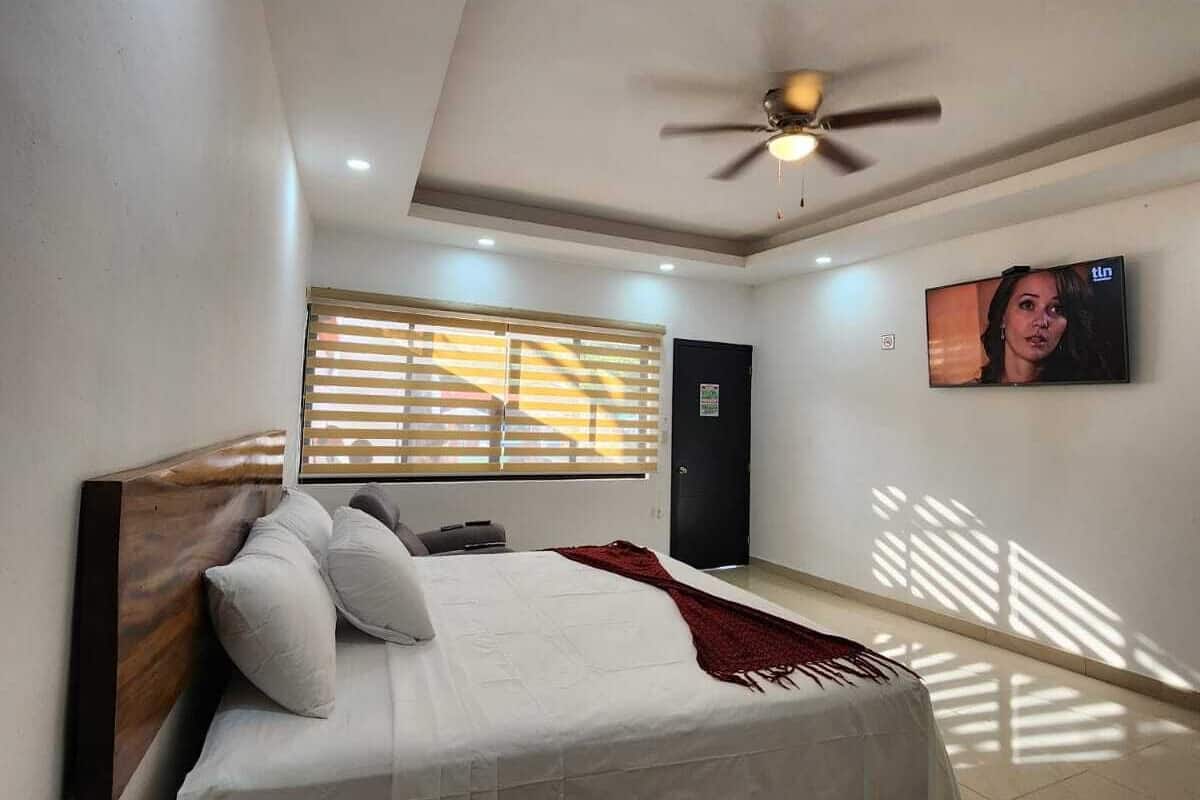 Ein Schlafzimmer mit Bett, Deckenventilator, Fernseher und einem Fenster mit Jalousien.