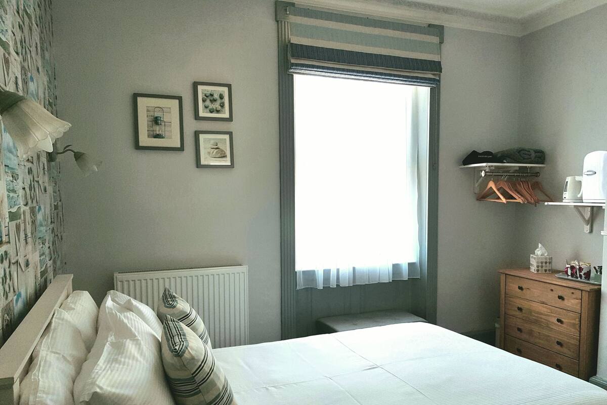 A bedroom with a bed, a radiator, a window with curtains, a wooden dresser, and wall-mounted shelves with items.