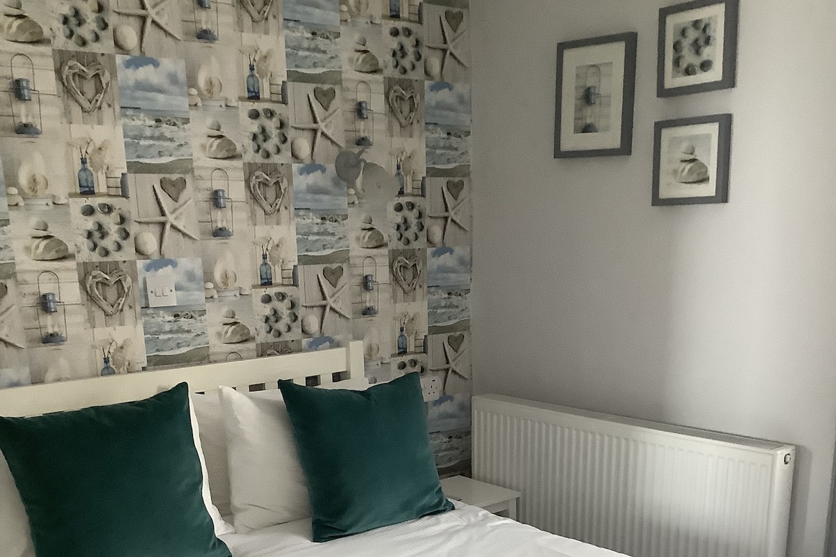 A neatly made bed with white linens and two teal pillows, a white headboard, and a wall decorated with various framed pictures and a collage of seashells and beach-themed items.