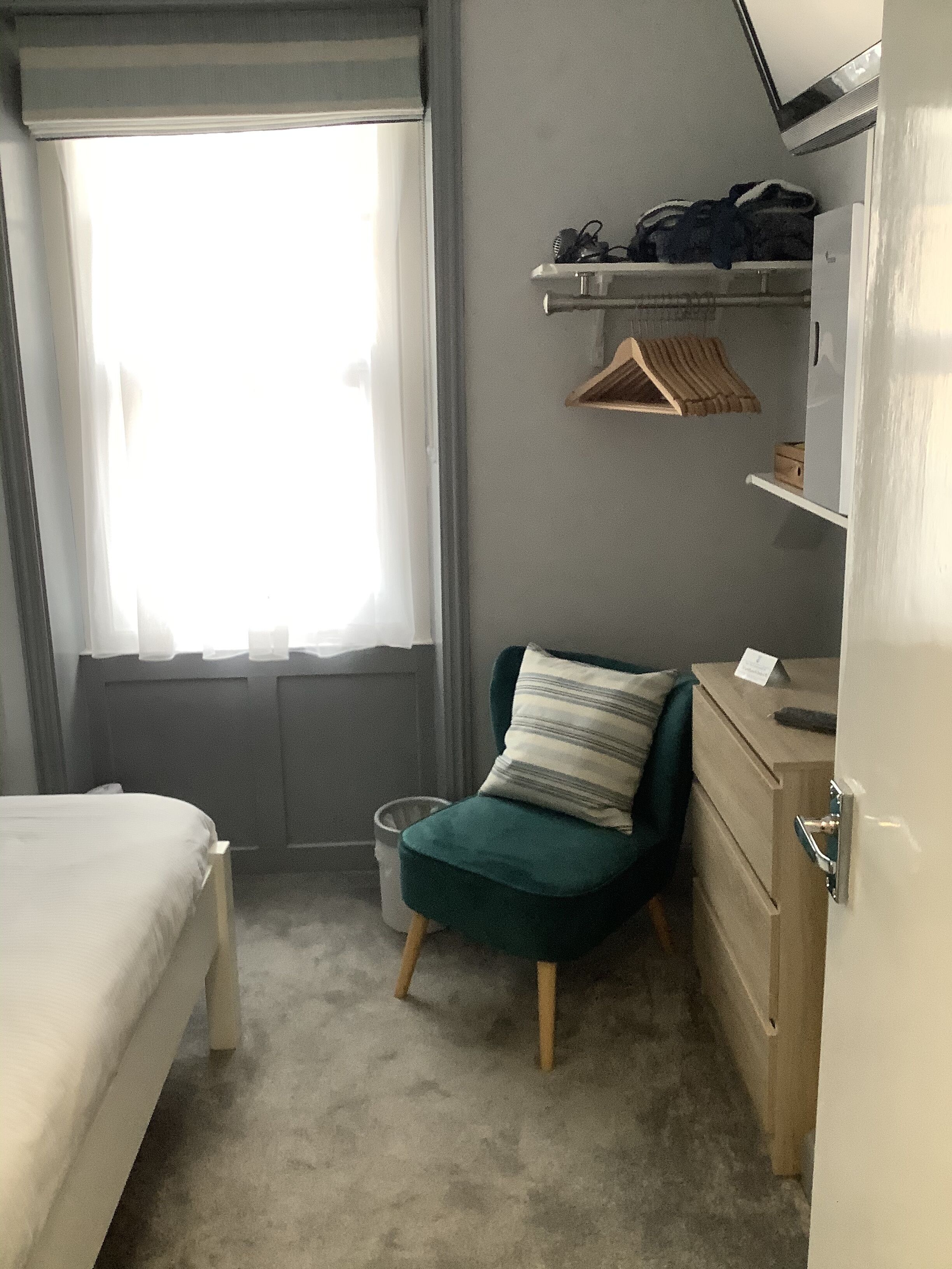 A small, compact room with a bed, a green chair with a striped pillow, a small desk with a mirror, and a window with sheer curtains.
