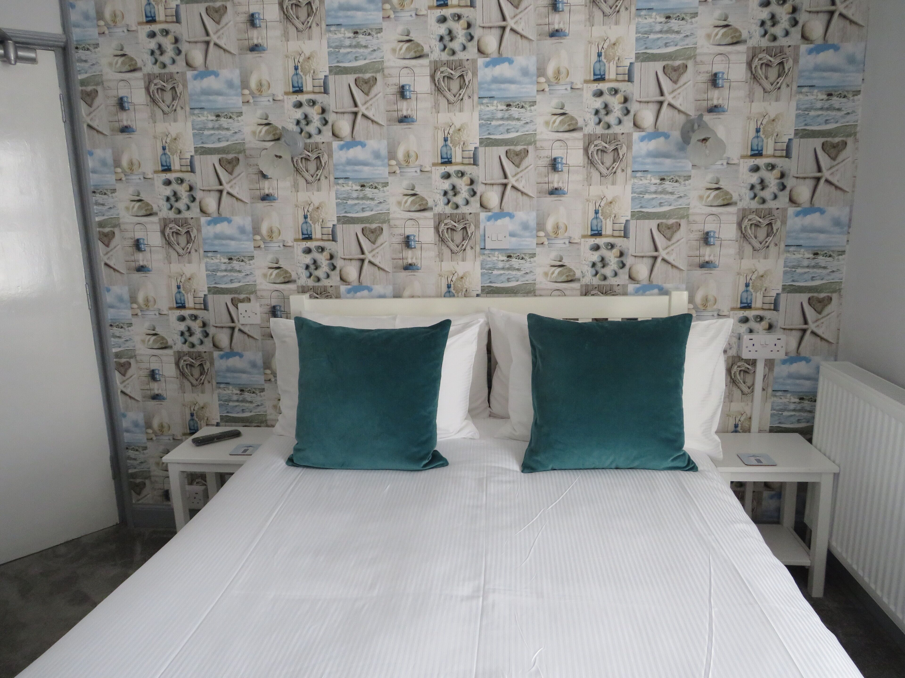 A bedroom with a bed, white bedding, teal pillows, and a wall decorated with nautical-themed tiles.