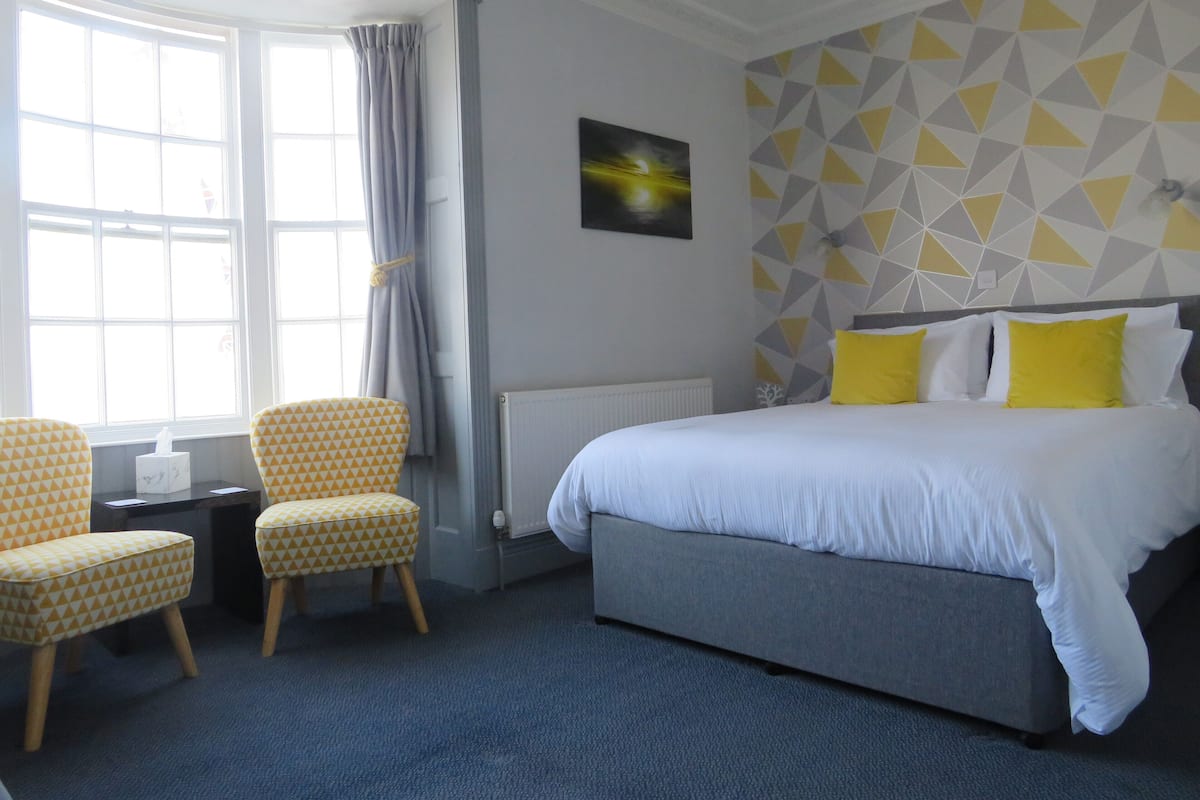 A hotel room with a bed, two patterned chairs, a table with a tissue box, a radiator, and a geometric-patterned wall.