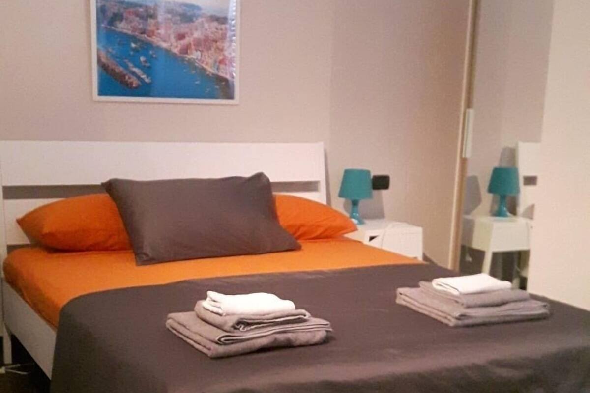 Une chambre à coucher moderne avec un lit, des oreillers, une serviette pliée, une table de chevet, une lampe et un tableau au mur.