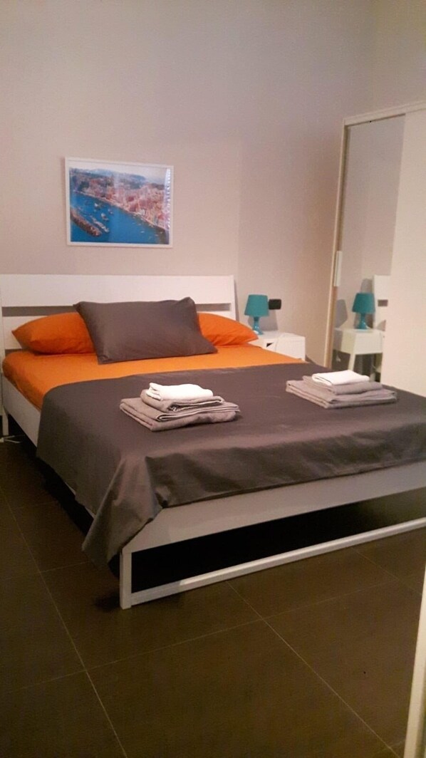 Un dormitorio moderno con cama, almohadas, una toalla doblada, una mesita de noche, una lámpara y un cuadro en la pared.
