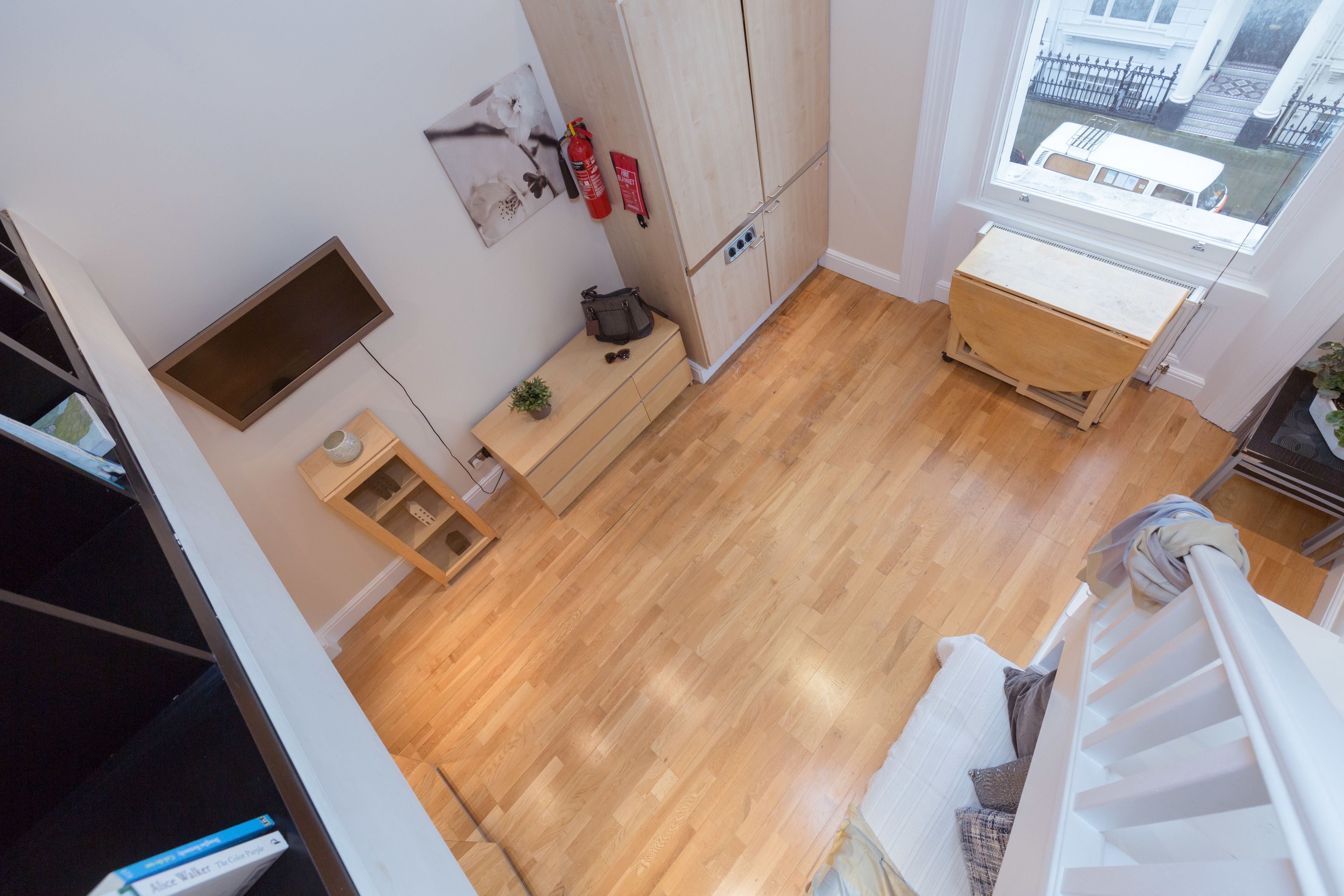 Quarto com piso de madeira, guarda-roupa branco, pequena mesa com planta e janela com vista para o exterior.