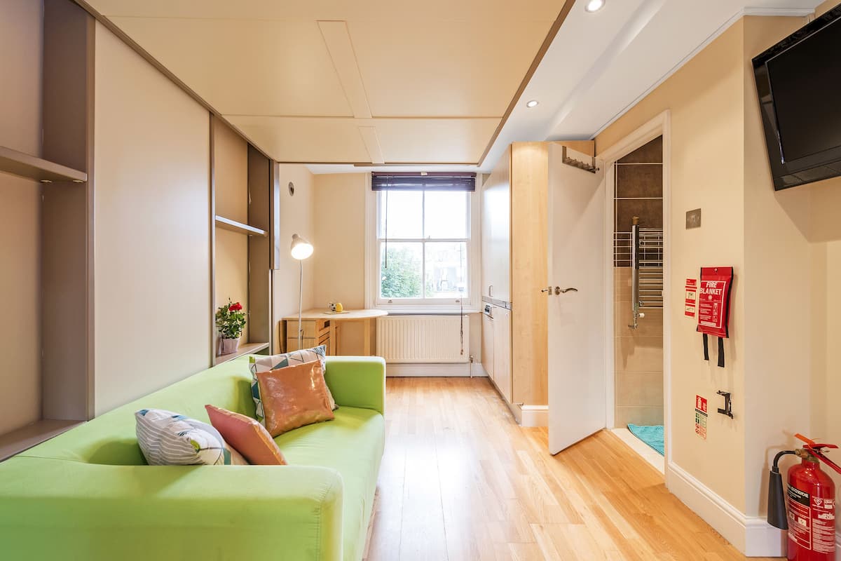 A compact room with a green sofa, a small table, a window, and a door leading to a bathroom.