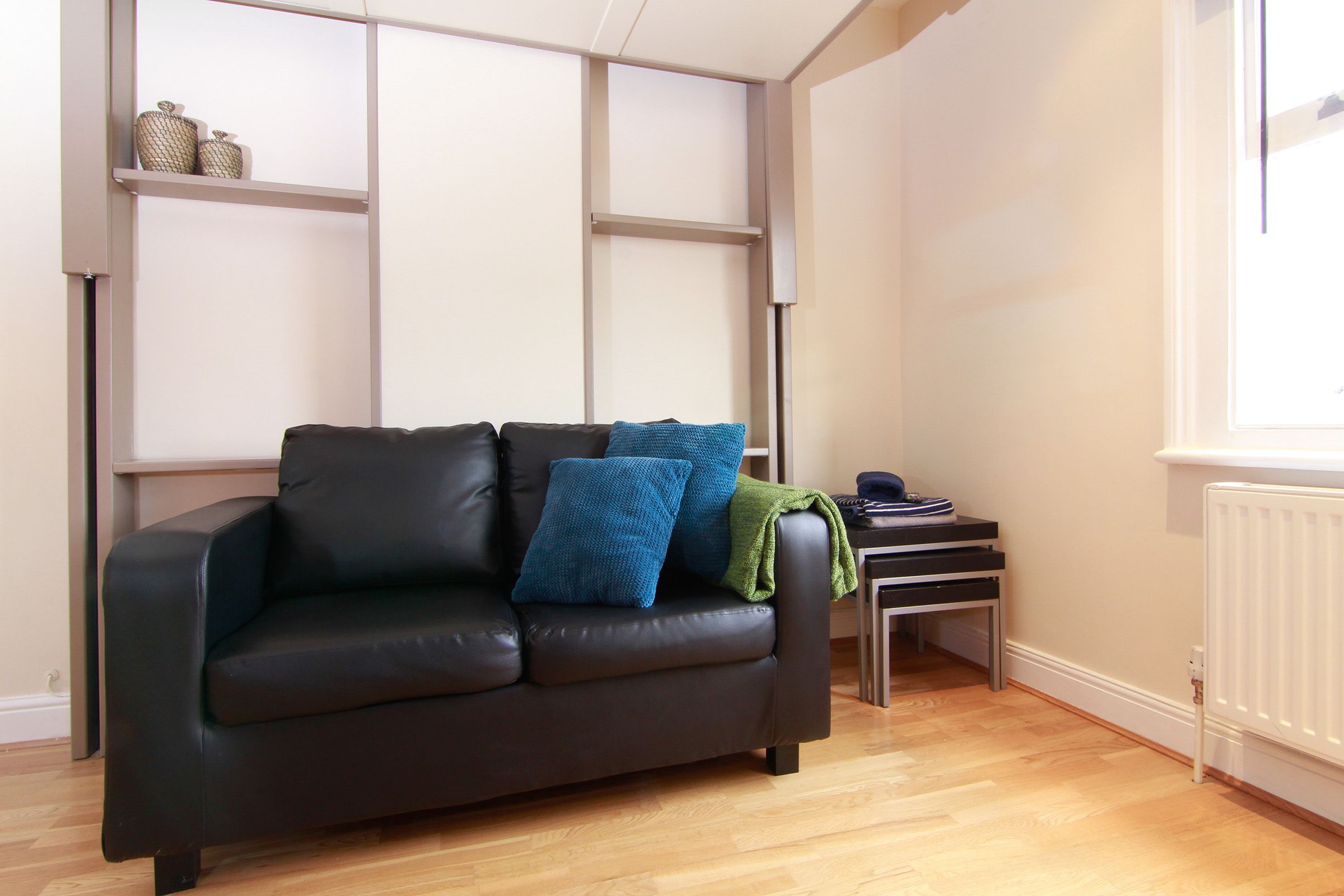 A modern living room with a black leather sofa, blue and green cushions, a small side table, and a radiator.